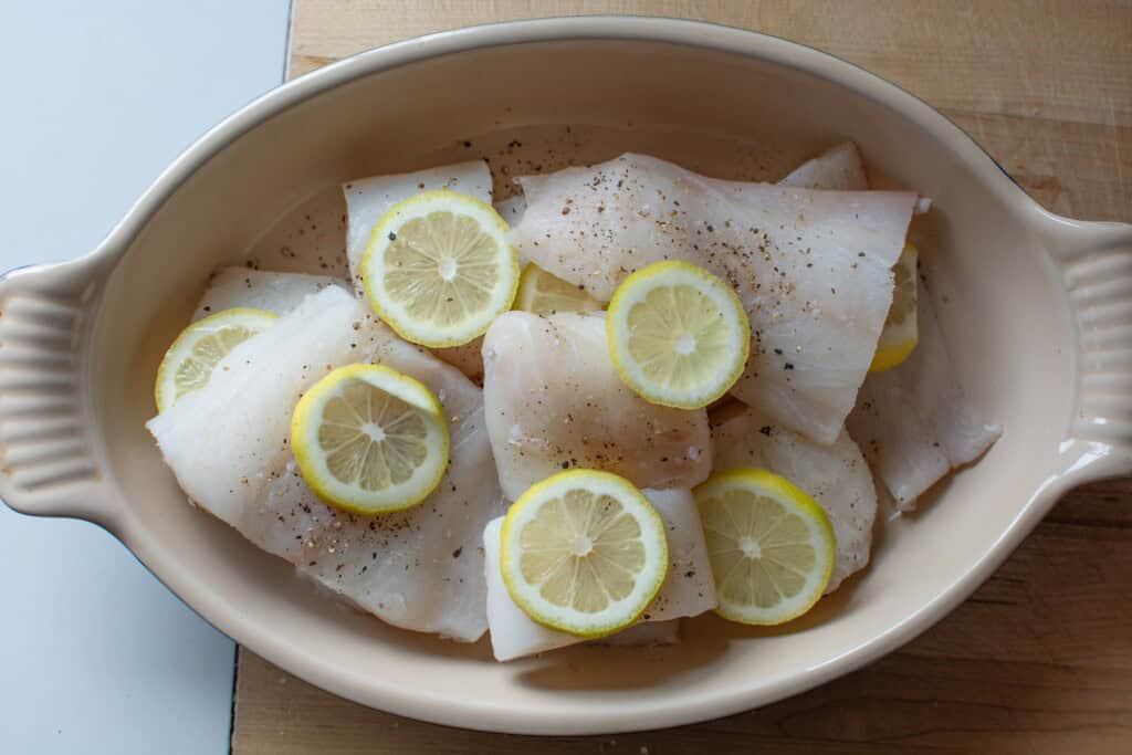 Raw cod fillets in a dish with slices of lemon on top. 