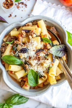 A bowl of pasta surrounded by red pepper flakes and basil leaves.
