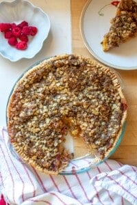 A peach and raspberry pie with crumb topping next to a bowl of raspberries.