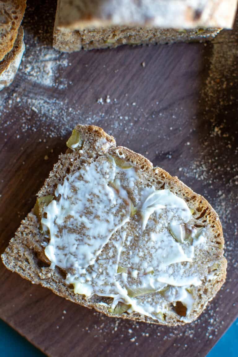 A slice of green olive bread slathered with butter on a wood cutting board.