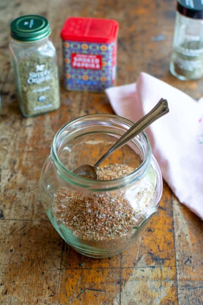 A jar of spice rub with the spices behind it on a wood table. 