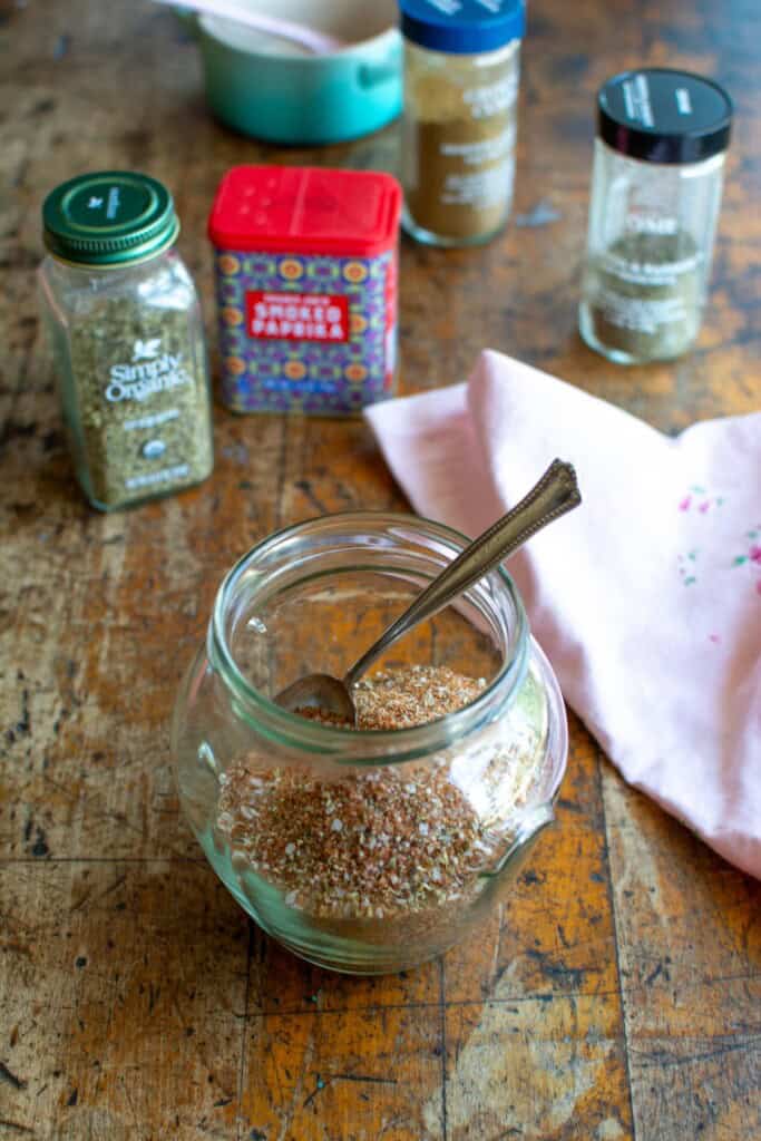 A jar of spice rub with all the spices behind it. 