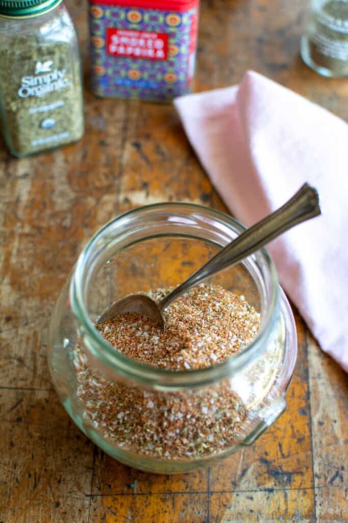 A jar of spice rub on a wood table with a spoon in the jar. 