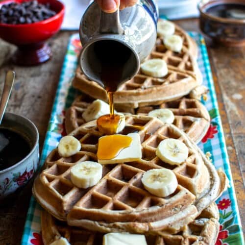 Waffles on a blue tray with a person pouring maple syrup all over them.