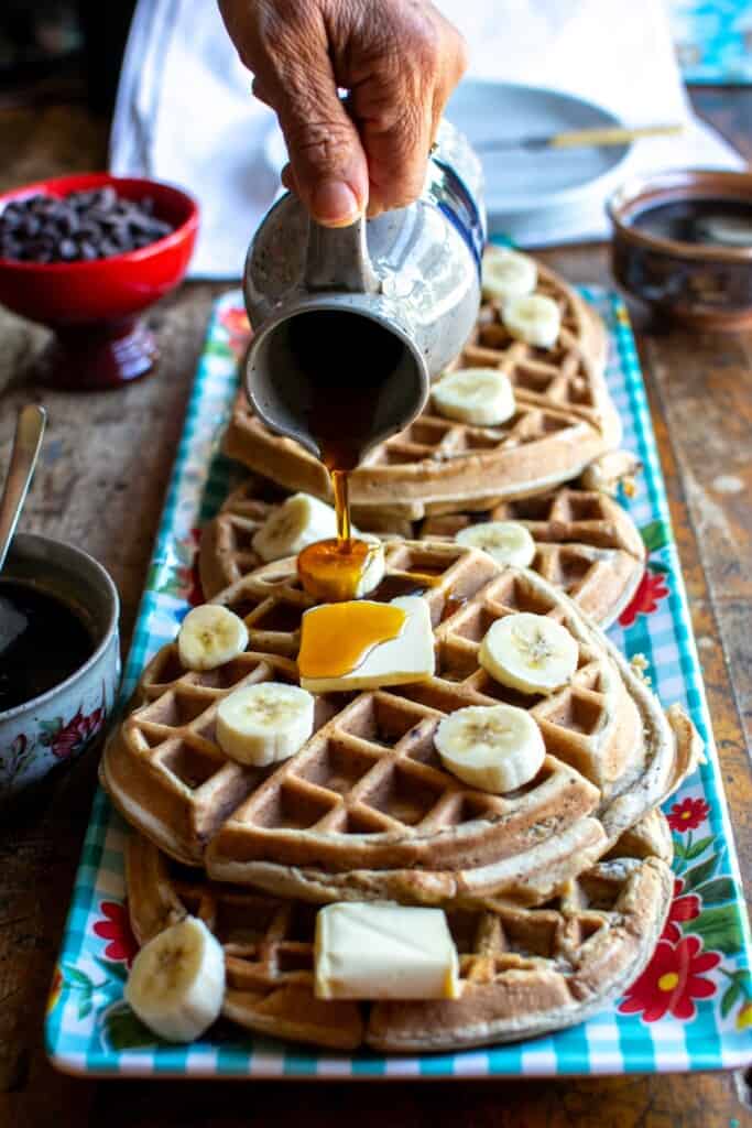 Waffles on a blue tray with a person pouring maple syrup all over them. 