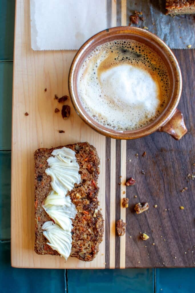 A piece of nutty apple bread with butter on it next to a cappuccino