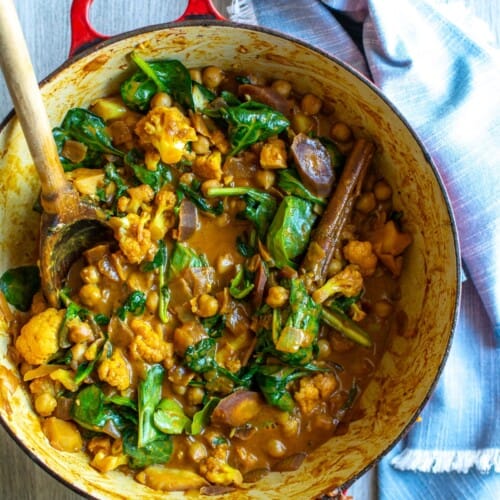 A red pot filled with vegetable curry with a wooden spoon in it.